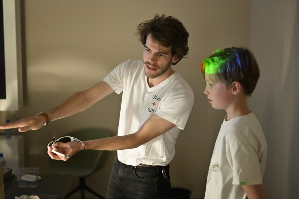 An SRON employee in white t-short explains the dispersion and polarimetrty of light to a boy. The boy has dispersed light sshining on his hair and colouring it in the colours of he rainbow.