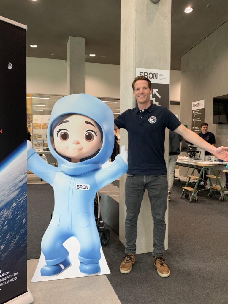 Een host bij de entree van de SRON open dag staat naast een levensgrote schattige masccotte die een cleanroompakje aan heeft.