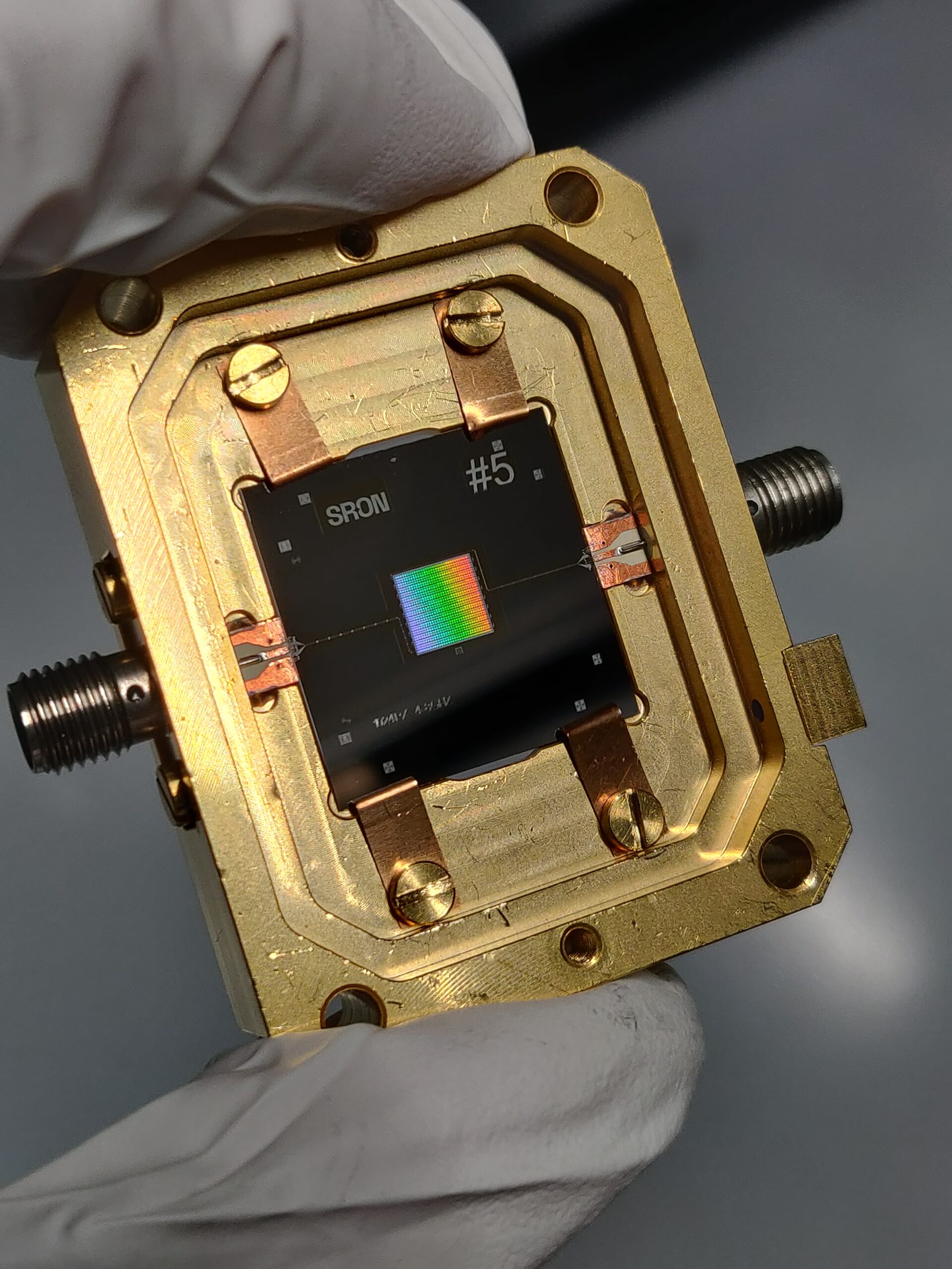 Tussen duim en wijsvinger wordt een KID array gehouden. De array schijnt in alle kleuren van de regenboog.